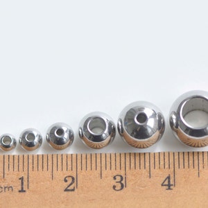 May include: A set of six silver metal beads of varying sizes, measured in centimeters on a ruler. The beads have a hole in the center.