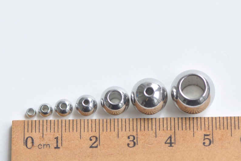 May include: A set of six silver metal beads of varying sizes, measured in centimeters on a ruler. The beads have a hole in the center.
