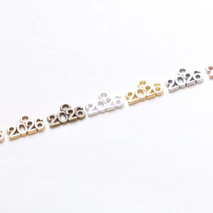 May include: A selection of metal charms in various metallic finishes, each displaying the year "2026". The charms are in rose gold, gold, bronze, silver, and other tones. Suitable for jewellery making or craft projects.