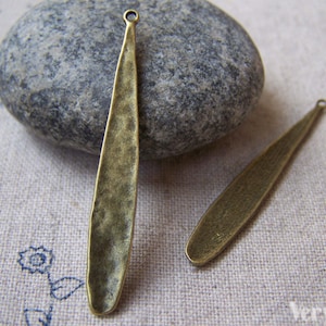 Op de afbeelding: Twee antieke goudkleurige metalen bedels in de vorm van lange, smalle bladeren met een gehamerde textuur. De bedels hebben een lus aan de bovenkant om aan sieraden of andere knutselwerkjes te bevestigen.
