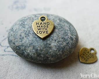 May include: Two small, heart-shaped charms with the words "HAND MADE WITH LOVE" engraved on them. The charms are a bronze colour and have a loop at the top for attaching to a necklace or bracelet. The charms are on a grey stone.