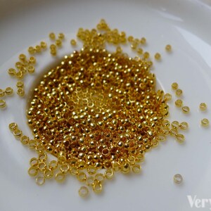 May include: A white ceramic dish filled with a large quantity of small, gold-colored metal jump rings. The jump rings are scattered around the dish, with some spilling over the edge.