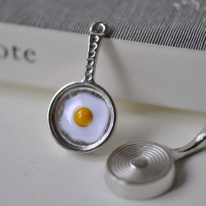 May include: Two silver charm pendants shaped like frying pans. One pan has a white egg with a yellow yolk inside. The other pan is plain.