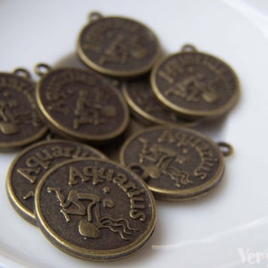 May include: A group of eight antique gold-toned metal charms featuring the astrological sign Aquarius. Each charm has a raised image of the Aquarius symbol and the word "Aquarius" written around the edge.