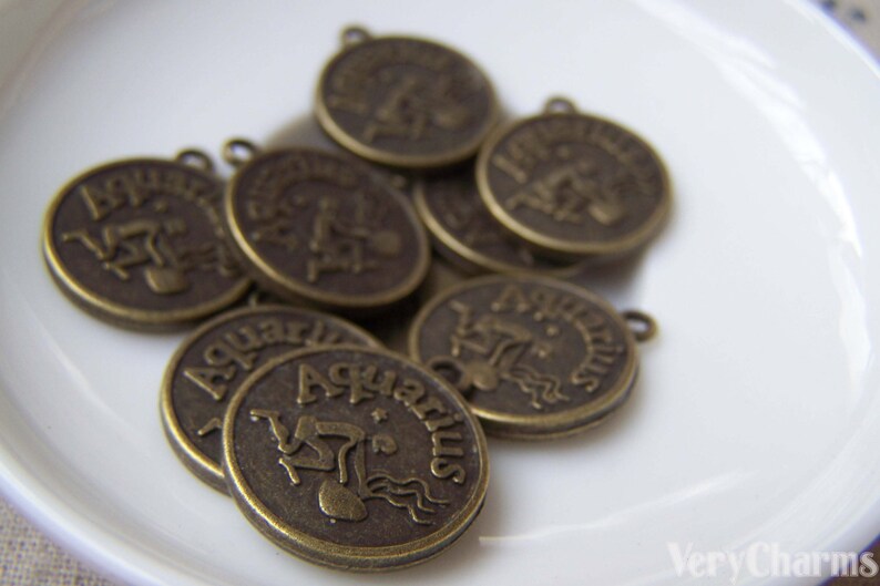 May include: A group of eight antique gold-toned metal charms featuring the astrological sign Aquarius. Each charm has a raised image of the Aquarius symbol and the word "Aquarius" written around the edge.