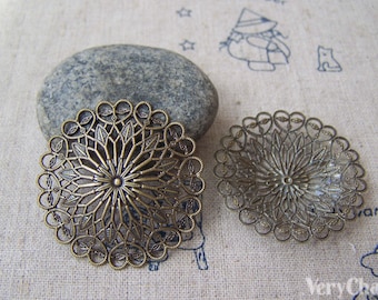 Puede incluir: Dos adornos redondos de filigrana de metal dorado antiguo con un diseño floral. El adorno más grande está sobre una roca gris.