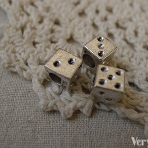 May include: Three silver dice charms with black dots. The dice are on a white crocheted surface.