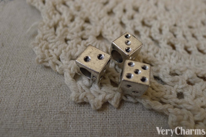 May include: Three silver dice charms with black dots. The dice are on a white crocheted surface.