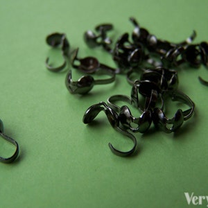 May include: A close-up of a pile of black metal jewelry findings. The findings are shaped like small hooks with a loop at the top. They are scattered on a green surface.