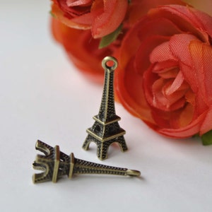 May include: Two small, gold-colored Eiffel Tower charms. The charms are on a white surface with a red rose in the background.