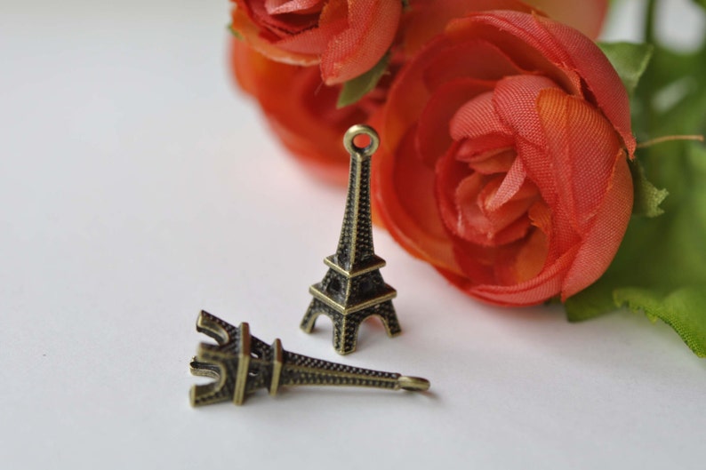 May include: Two small, gold-colored Eiffel Tower charms. The charms are on a white surface with a red rose in the background.