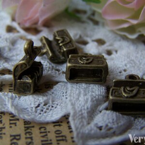 May include: Five antique gold-toned metal charms shaped like small boxes with intricate designs. The charms are on a white lace background.