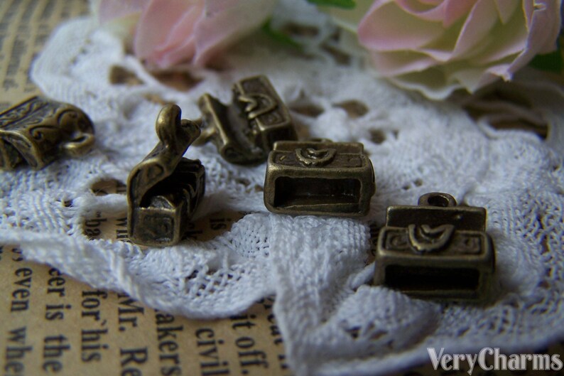 May include: Five antique gold-toned metal charms shaped like small boxes with intricate designs. The charms are on a white lace background.