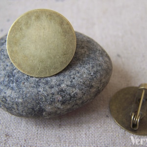 Peut inclure: Deux disques métalliques ronds, vierges, de couleur laiton. Un disque est posé sur un rocher gris. L'autre disque est muni d'un fermoir à épingle.
