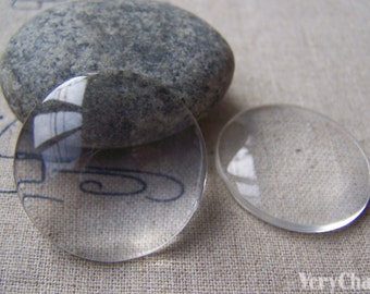 May include: Two clear glass cabochons, one larger than the other, on a light brown fabric surface.