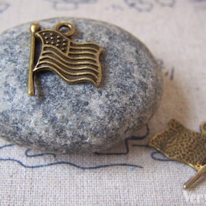 Peut inclure: Deux breloques en métal doré vieilli en forme de drapeau américain. Les drapeaux sont sur une pierre grise et un fond en tissu beige.