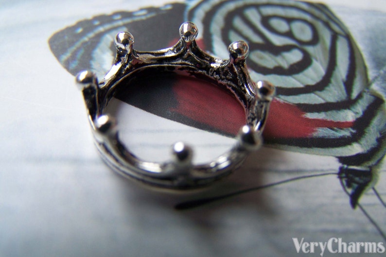 Puede incluir: Un charm de corona plateado con un dise&ntilde;o detallado. La corona es redonda y tiene cinco puntas. El charm est&aacute; sobre un fondo blanco.