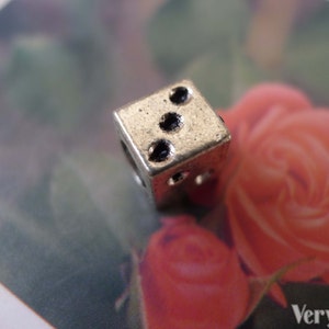 May include: A silver charm in the shape of a dice with two dots on one side. The charm is on a white background with a blurry image of a pink rose.