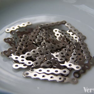 Puede incluir: Un montón de eslabones de cadena de metal plateados en un plato de cerámica blanco. Los eslabones son pequeños y tienen un diseño decorativo.