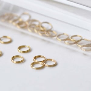 May include: A glass jar filled with gold jump rings, with a few jump rings scattered on a white surface.