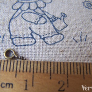 May include: A close-up of a small, gold-colored metal screw eye with a loop on one end. The screw eye is measured against a ruler, showing it is just over 1 cm long.