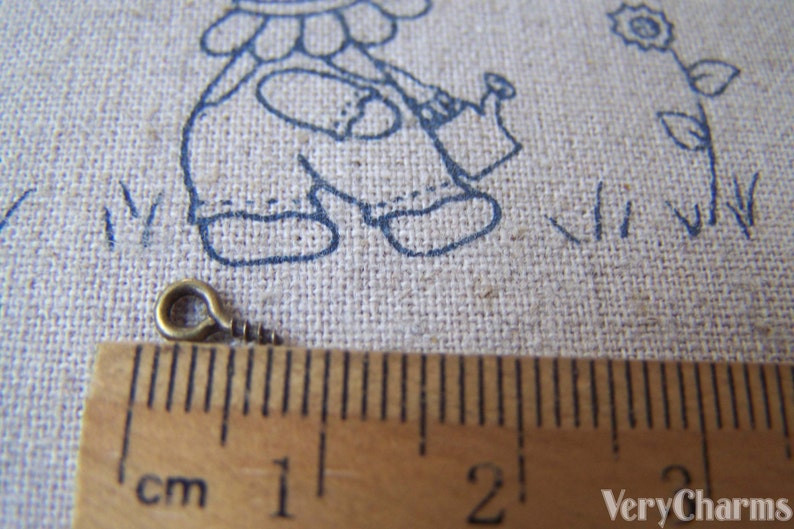 May include: A close-up of a small, gold-colored metal screw eye with a loop on one end. The screw eye is measured against a ruler, showing it is just over 1 cm long.