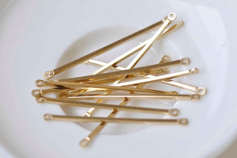 May include: A group of gold-colored metal jump rings with a loop on each end. The rings are arranged in a pile on a white surface.