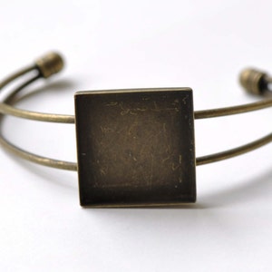Puede incluir: Un brazalete de doble cadena de bronce con un bisel cuadrado para un camafeo u otro adorno.