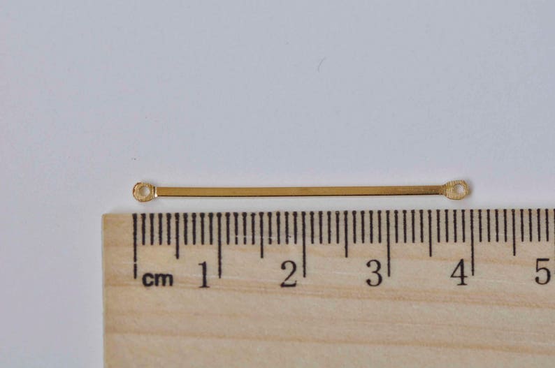 May include: A gold-colored metal bar with two small loops at each end. The bar is approximately 3 cm long.