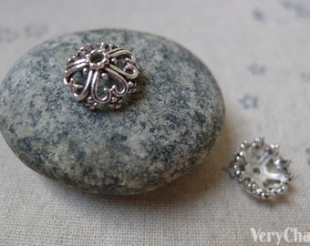 May include: Two silver metal bead caps with a decorative, ornate design. The caps are on a gray stone and a white fabric background.