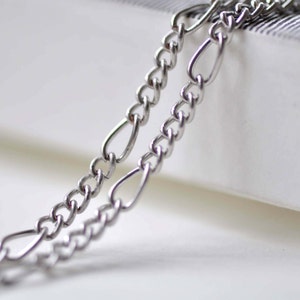 May include: Two silver chains, one thicker than the other, are linked together. The chains are lying on a white surface.