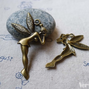 Op de afbeelding: Twee antieke goudkleurige metalen fee-charms. Een charme is een fee met uitgespreide vleugels, de andere is een fee met gevouwen vleugels en haar hoofd gebogen.