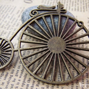 Könnte beinhalten: Ein bronzefarbener Metallanhänger in Form eines Vintage-Fahrrads mit einem großen, Speichenrad. Der Anhänger hat ein detailliertes Design und ist perfekt, um Schmuck oder Bastelarbeiten einen Hauch von Steampunk-Stil zu verleihen.