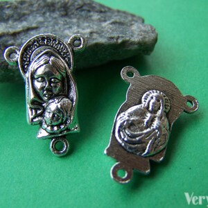 May include: Two silver-toned metal charms featuring the image of the Virgin Mary and baby Jesus. The charms have a loop on each side for attaching to a bracelet or necklace.