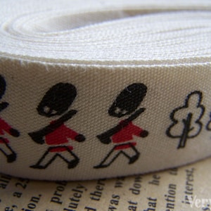 May include: White cotton ribbon with a repeating pattern of red and black stick figures wearing hats and carrying rifles, and two black trees.