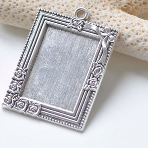 May include: Silver-toned metal picture frame pendant with a decorative floral design. The frame has a raised edge and a smooth, textured surface.