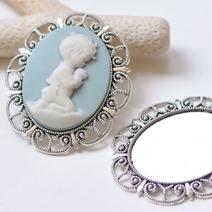 Op de afbeelding: Twee zilverkleurige metalen cameo-lijsten met sierlijke filigraanontwerpen. Een lijst heeft een blauwe cameo met een wit porseleinen beeldje van een kind dat bidt. De andere lijst is leeg.
