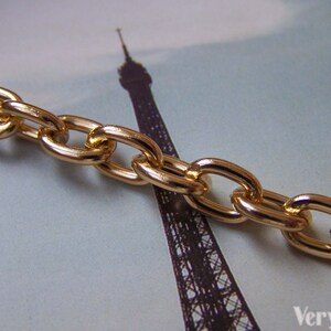 May include: Gold chain link with oval shaped links. The chain is lying on a surface with a blurred image of the Eiffel Tower in the background.