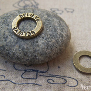 Könnte beinhalten: Ein goldfarbener Metall-Anhänger mit dem Wort "BELIEVE" eingraviert um den Rand. Der Anhänger liegt auf einem grauen Stein.