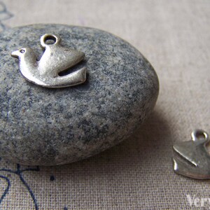 May include: Two silver dove charms, one on a gray stone and one on a beige fabric background. The dove charms are simple and elegant, with a classic design.
