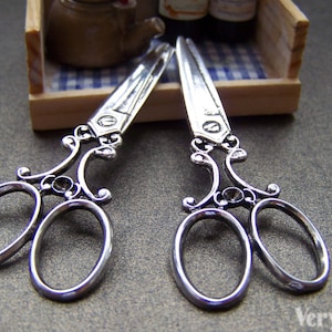 May include: Two silver-toned decorative scissors with ornate handles. The scissors are open and the blades are pointed.