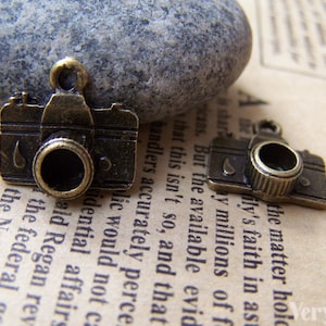 May include: Two antique gold-toned camera charms. The charms are detailed with a lens and a textured surface.