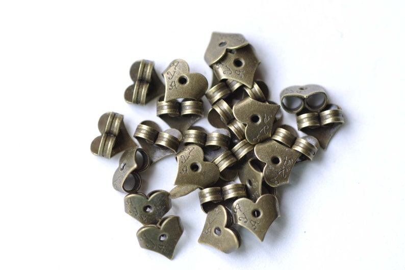 Op de afbeelding: Een stapel antieke gouden hartvormige metalen bedels met een klein gaatje in het midden van elk hart. De bedels hebben een decoratief ontwerp en de tekst "24" is op elke bedel gegraveerd.
