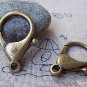 Puede incluir: Dos cierres de langosta de metal de color dorado antiguo. Los cierres están abiertos y listos para ser unidos a una cadena u otras joyas.