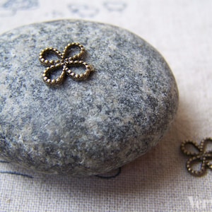 May include: Two small, bronze, decorative metal flower charms. The charms are on a gray stone and a white fabric surface.