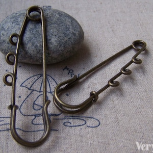 May include: Two antique bronze safety pins, one with a decorative coil design and the other with multiple loops. The pins are lying on a beige fabric surface.