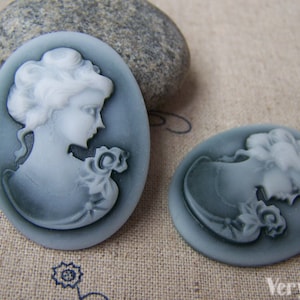 May include: Two oval-shaped cameo pendants featuring a white cameo image of a woman's profile on a grey background.