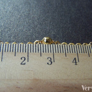 May include: Close-up of a gold-colored necklace with small, round beads spaced along the chain. The necklace is measured against a wooden ruler, showing centimeters. The ruler is marked from 1 to 5 cm.