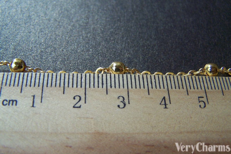 May include: Close-up of a gold-colored necklace with small, round beads spaced along the chain. The necklace is measured against a wooden ruler, showing centimeters. The ruler is marked from 1 to 5 cm.