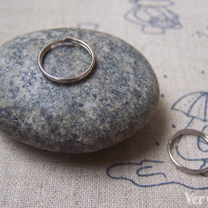 May include: Two silver jump rings, one on a gray stone and one on a white fabric background with a drawing of a person holding an umbrella.